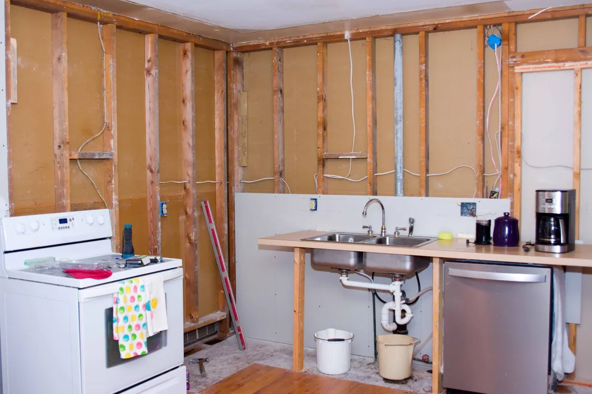 Kitchen undergoing renovation: exposed studs, appliances (stove, sink, dishwasher) installed, unfinished walls.