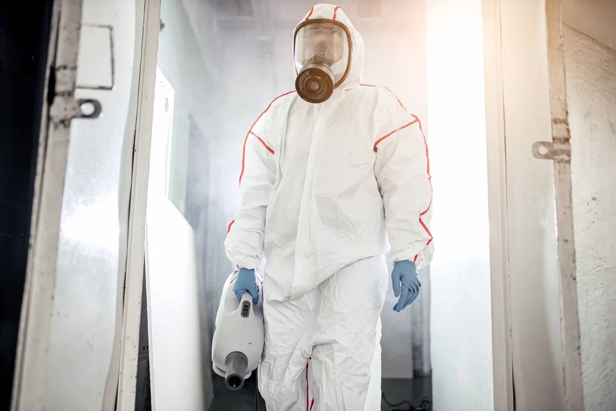 A person in a protective suit and respirator steps through a doorway carrying a sealed bag.