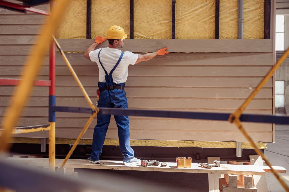 Construction worker installing siding on a building, wearing a hard hat and safety gloves.