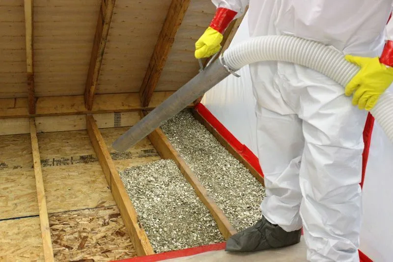 A worker in a white suit and gloves vacuums vermiculite from between attic joists, behind a polyethylene containment.