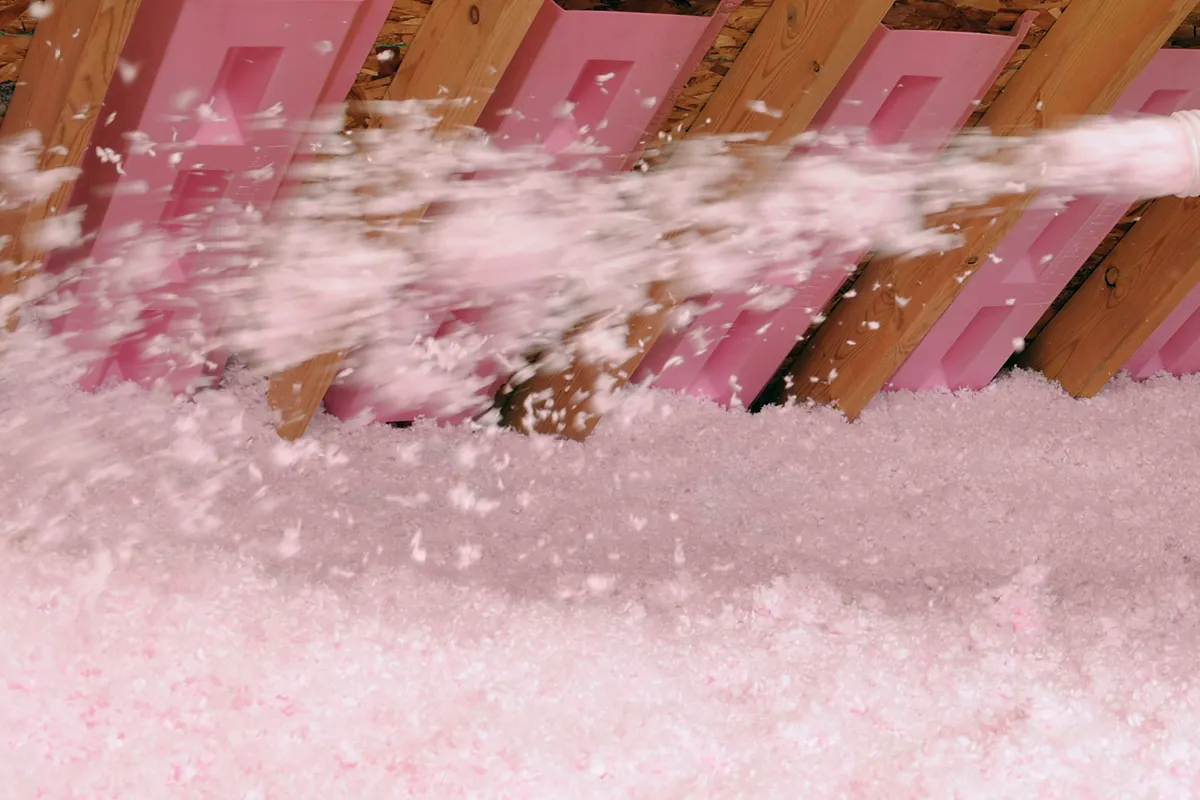 Pink insulation being blown into an attic with exposed wooden rafters and pink foam boards.